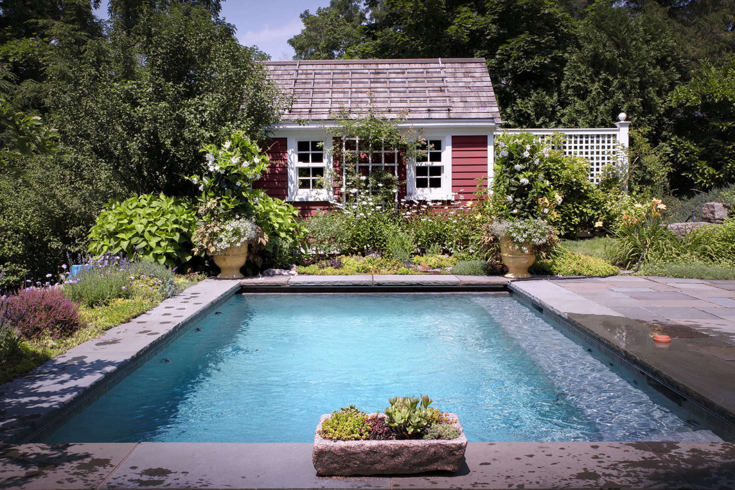 Surrounded by abundant perennials such as Shasta daisies, as well as terra cotta pots planted with vibrant annuals, the rose-covered pool house enhances the cottage feel.